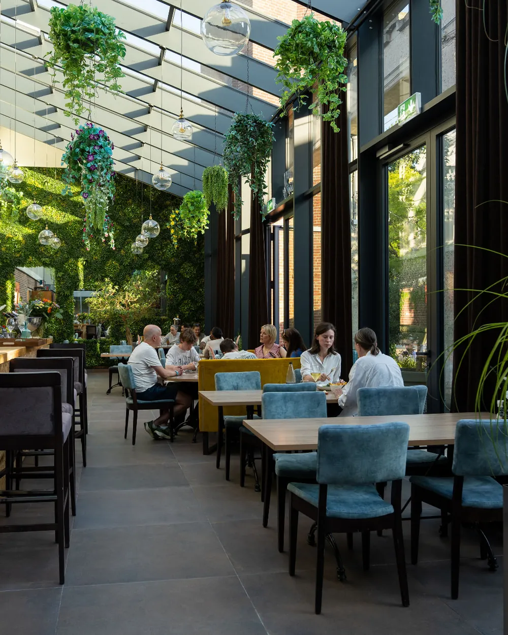 Gasten dineren in de lichte serre van Brasserie Lenny in Amsterdam Oost, met groene wand, hangplanten en de bar op de achtergrond